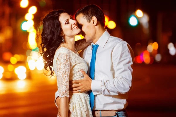 Elegant dressed man is tenderly embracing happy smiling woman at bright night street lights background.