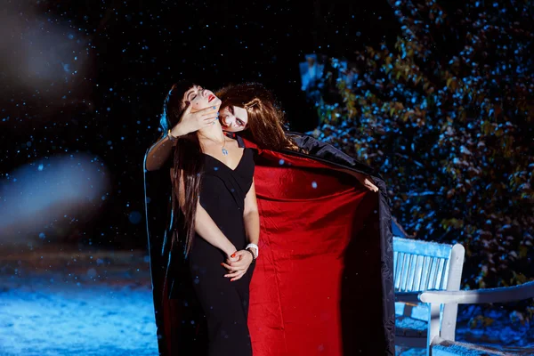 Beautiful female vampire in long pallium is biting her victim at blue light cold winter evening background.