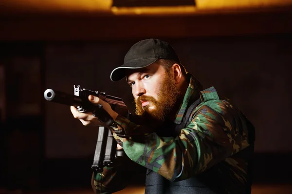 Terrorist in camouflage with red beard and cruel face is holding rifle at indoors background.