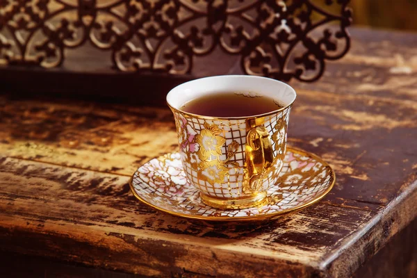 Closeup image of vintage porcelain tea cup on saucer with golden floral pattern at wooden desk background.