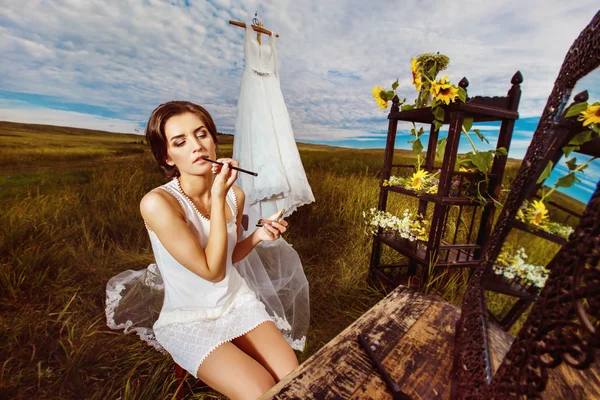 Portrait of beautiful bride making makeup at vintage mirror on commode outdoors at summer field background.