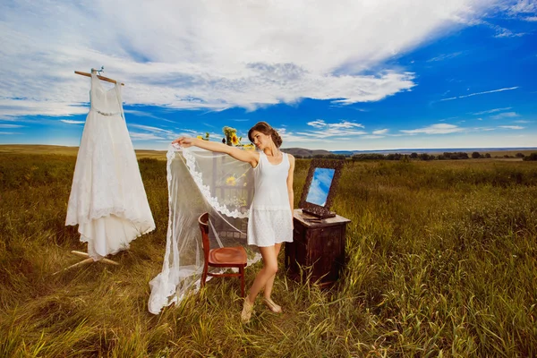 Concept of bride morning. Beautiful bride is standing outdoors holding veil at amazing summer rural landscape field background with sunflowers decoration and wedding dress on rack.