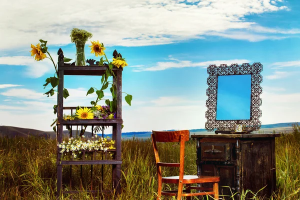 Beautiful rural landscape of field with vintage furniture decorated with wild flowers at blue summer sky background.