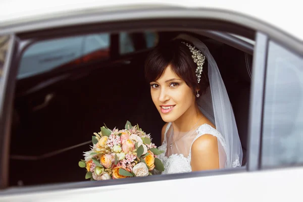 Gorgeous bride with beautiful smile is sitting in wedding car and holding summer flowers bouquet.