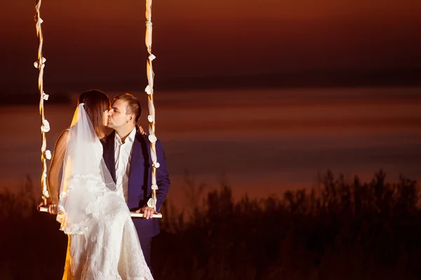 Beautiful wedding couple is kissing on swing at golden sea sunset background.