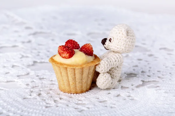 Small knitted teddy bear sitting near mini tartlet.