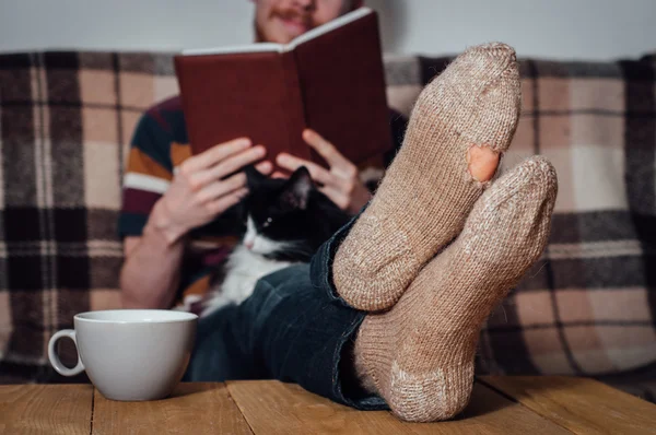 Young man reading book on couch with cat in holey socks