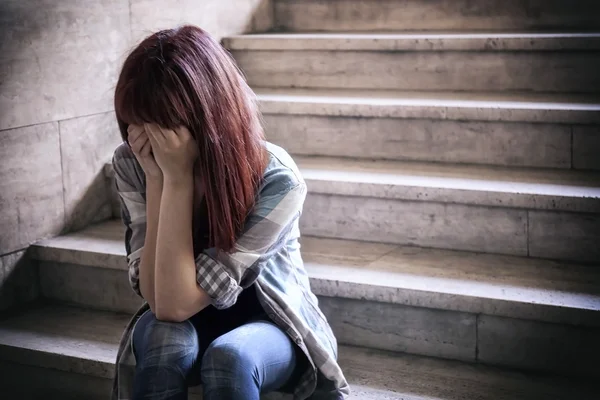 Depressed girl in adolescent crisis sitting on the steps of a basement, covers her face with her hands. A natural light penetrates from above.