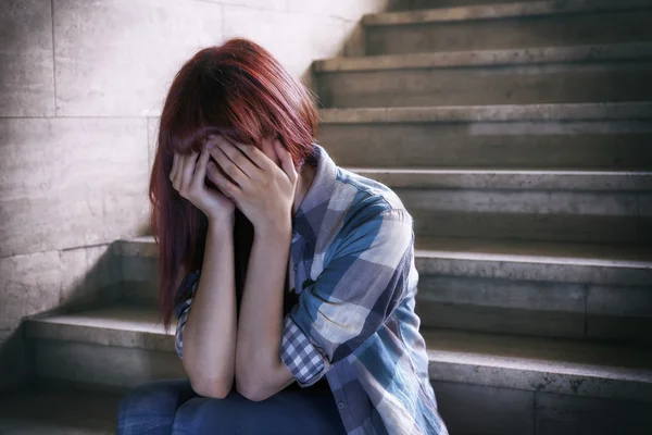 Depressed girl in adolescent crisis sitting on the steps of a basement, covers her face with her hands. A natural light penetrates from above.