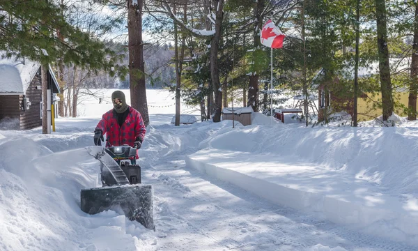 A man in work clothes, hat and gloves, operating a snow thrower / blower on a winter day in Canada.