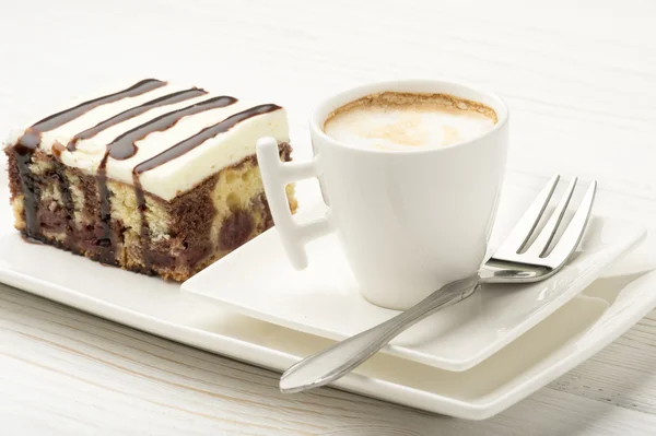 A piece of cake with cherry and chocolate and a cup of coffee on the white wooden background.