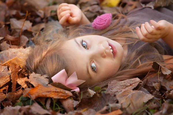Beautiful little girl with brown hair laying on the ground covered with leaves in fall