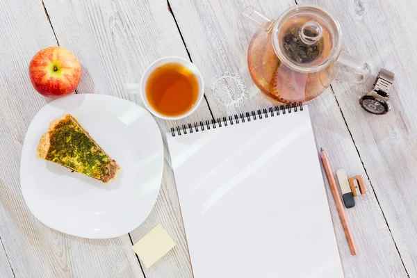 Drawing-pad, cup of tea, chicken pie, apple, glass teapot and watch on wooden table