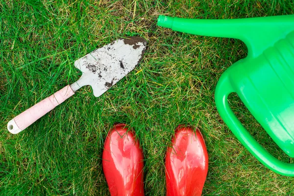 Gardening tools, red garden shoes, small spade,  watering can on the grass,  close up.