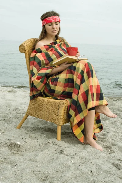 Hipster girl reading a book on the beach. Beautiful young hipster woman with book, blanket and cup of tea.