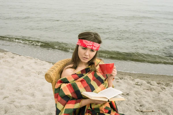 Hipster girl reading a book on the beach. Beautiful young hipster woman with book, blanket and cup of tea.