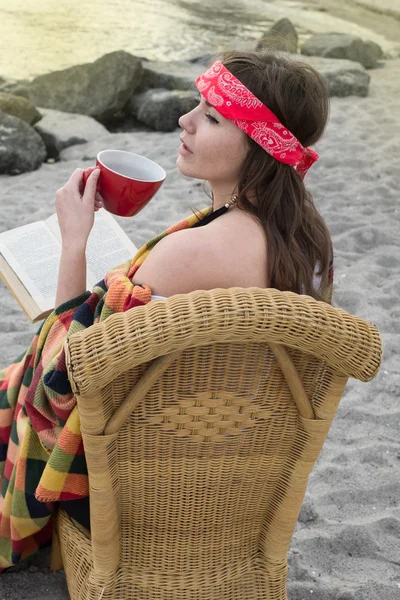 Hipster girl reading a book on the beach. Beautiful young hipster woman with book, blanket and cup of tea.