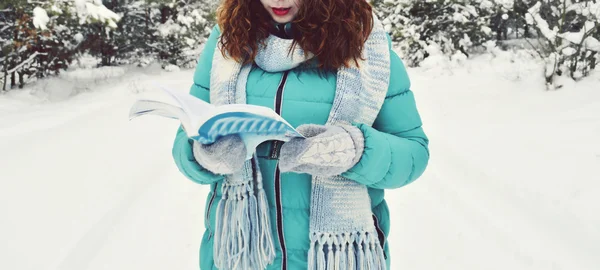 Dark-haired girl dressed in a turquoise jacket,bodily woolly hat, and a light blue scarf,reading a book in the Park,drinking tea,coffee,cocoa