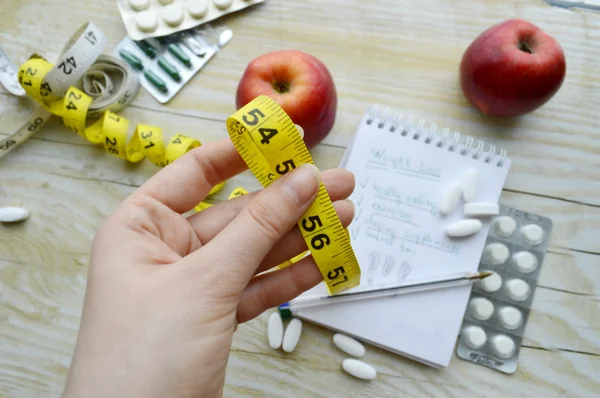 Female hand with measuring tape, pills, apples and notebook