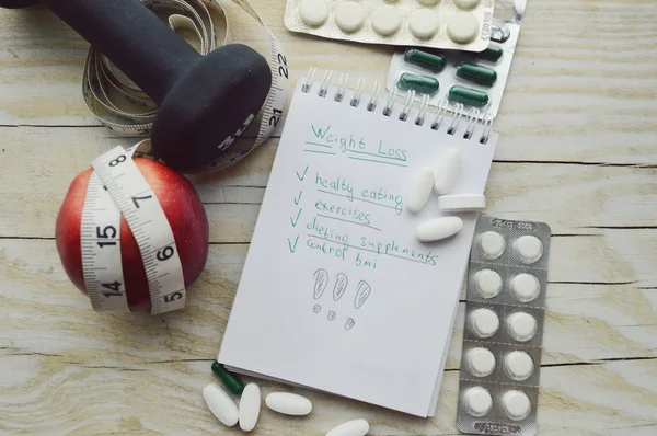 Notebook with pills, apple, measuring tape and dumbbell