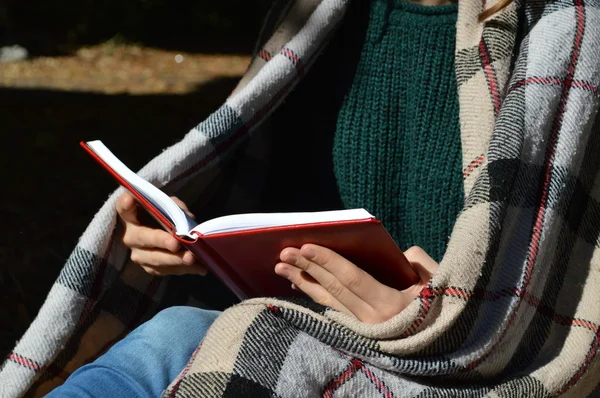 A young,beautiful girl wrapped in a warm plaid blanket drinking hot tea and reading a book in the Park