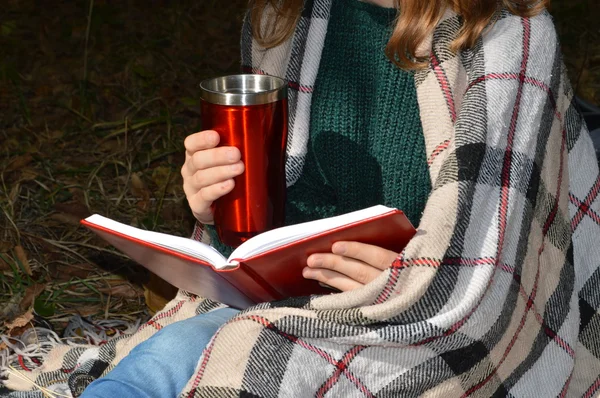 A young,beautiful girl wrapped in a warm plaid blanket drinking hot tea and reading a book in the Park