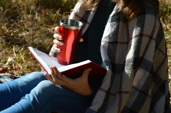 A young,beautiful girl wrapped in a warm plaid blanket drinking hot tea and reading a book in the Park