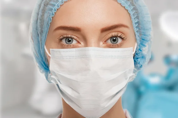 Close-up portrait of serious nurse or doctor in white mask. Looking confident and professional. Operation in the background