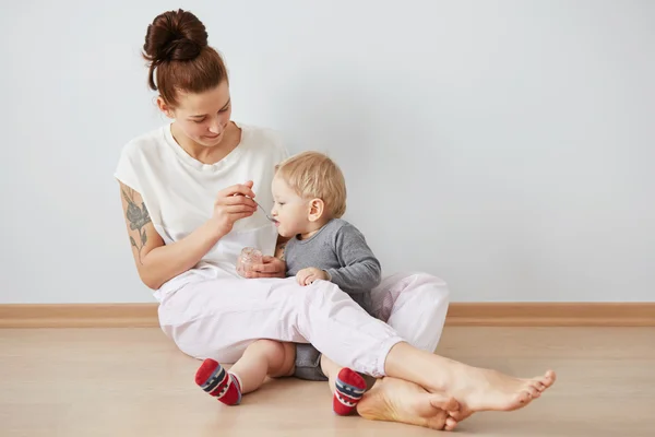 Young attractive mother with her one years old little son dressed in pajamas  giving him his first fruit smoothies food in the bedroom at the weekend together, lazy morning. Selective focus