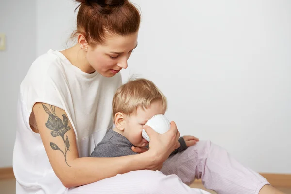 Young mother with her one years old little son dressed in pajamas  gives to drink water from cup in the bedroom at the weekend together, lazy morning, warm and cozy scene. Selective focus.