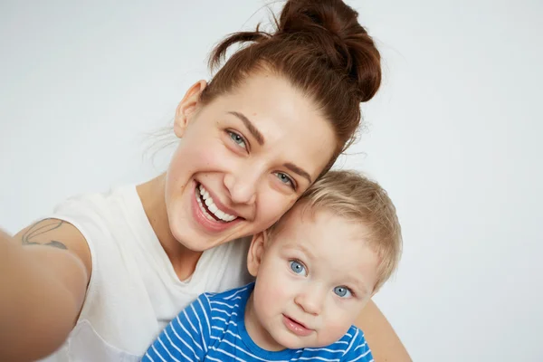 Young mother with her one years old little son dressed in pajamas are posing. Mom with son taking selfie on her smartphone or action camera in the bedroom.  Selective focus. Casual lifestyle photo.