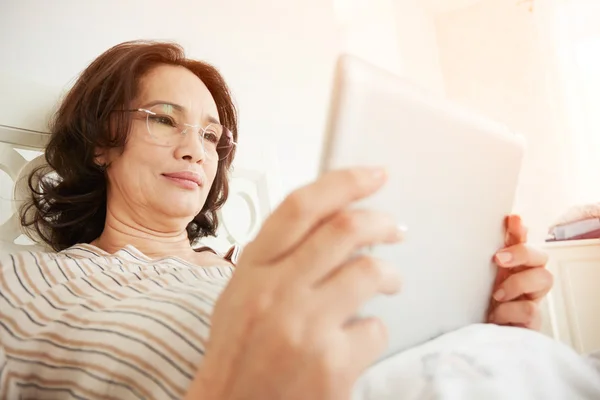 Smiling mature woman in glasses using digital tablet pc lying on her bed in a bedroom. Flare sun light