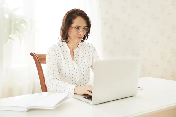 Beautiful asian mature female using portable laptop computer while work at compilation of business charts, woman sitting at the table with open net-book in office or home interior.