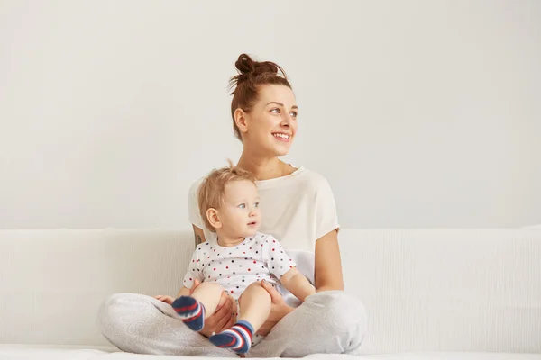 Young mother with her one years old little son dressed in pajamas are relaxing and playing in the bedroom at the weekend together, lazy morning, warm and cozy scene. Selective focus.