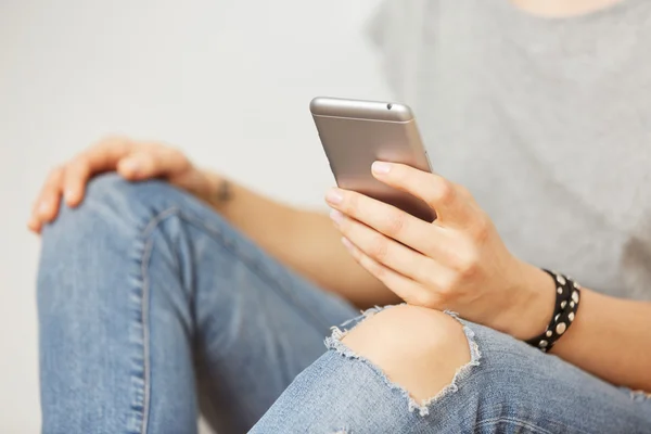 Beautiful hipster woman checking email via mobile phone while grey wall, charming female holding cell telephone in hands while waiting for friends.