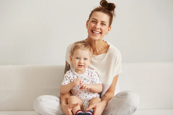 Charming brunette woman playing with her baby while sitting on a bed in her apartment at the weekend together, lazy morning, warm and cozy scene. Selective focus
