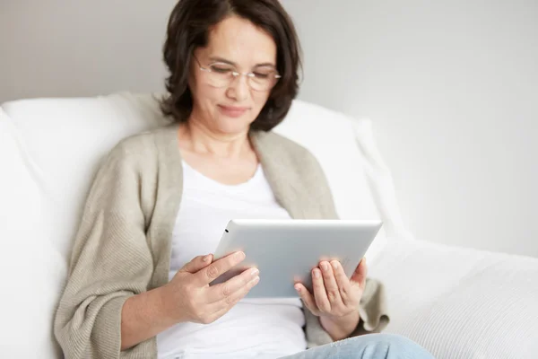 Middle-aged woman using touch pad reading a message, email or information on her tablet computer sits on a couch at home. Smiling senior woman at home connected on internet.