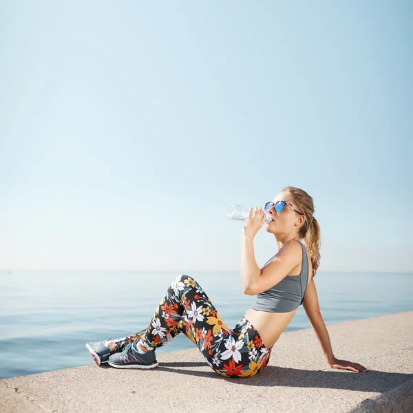 Muscular young female fitness blonde woman drinking water from bottle after running at beach. Woman sport runner resting taking a break with water bottle drink outside after training. Big size photo.