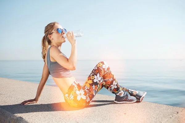 Muscular young female fitness blonde woman drinking water from bottle after running at beach. Woman sport runner resting taking a break with water bottle drink outside after training.