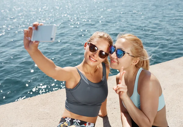 Happy fitness selfie blonde girls smiling and taking self portrait photograph with smart phone after running exercise workout on beach.  Healthy lifestyle with fit Asian and Caucasian women.