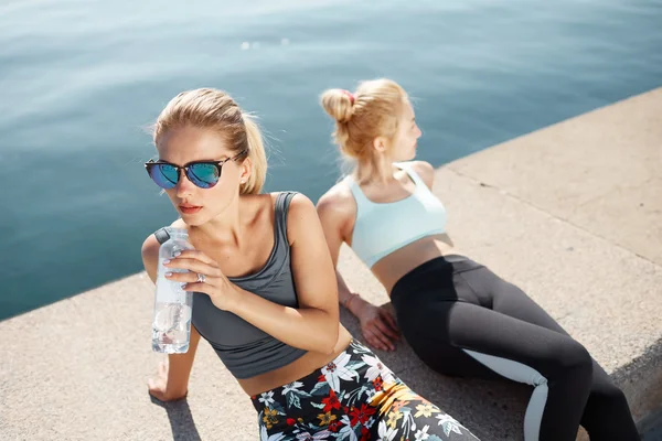 Two young women athlete having break after running exercise. Athletic females resting on the beach after physical exercises. Blonde active girls enjoying break during running training outdoors on sunn