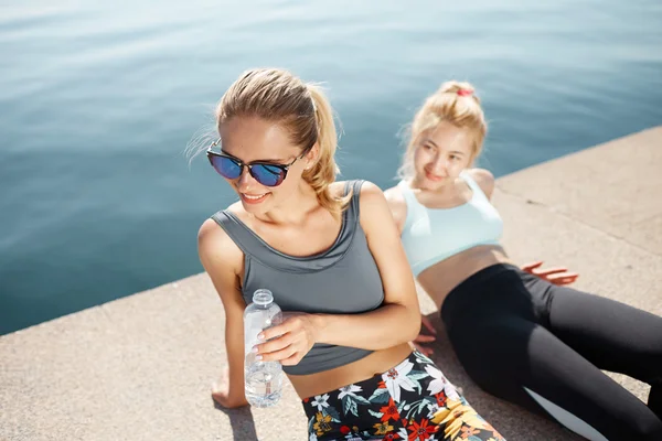 Portrait of two young fitness girls relaxing after training outdoors. Attractive fit running partners sitting on the beach after successful urban outdoor workout. Female sporty friends portrait