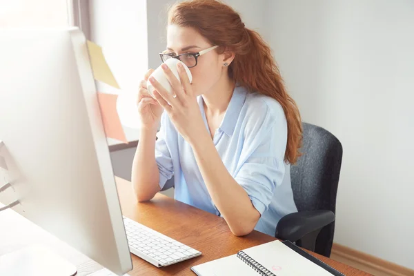 Young businesswoman reading something on the computer while enjoying coffee during lunch break in the office. Thoughtful female freelancer drinking coffee while waiting answer on her e-mail