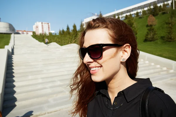 Close-up lifestyle outdoor portrait of pretty redhead hipster girl in sunglasses laughing and posing against urban background. Attractive student girl enjoying sunny day while having walk in the park