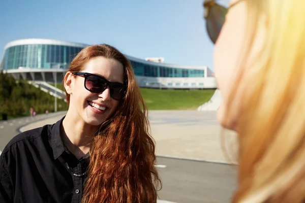 Young redhead woman in sunglasses posing against urban background. Outdoor portrait of two student girls meeting in the park, having a walk in the morning before classes at university. Selective focus