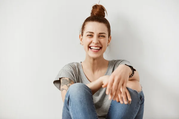 Close up portrait of young Caucasian woman in denim pants and gray T-shirt posing while seating on the floor with hands on her knees with happy expression enjoying free time at home. Selective focus