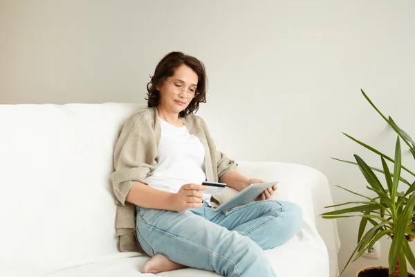 Smiling senior woman holding credit card and digital tablet, paying online for utility bills, sitting on white sofa. Mature woman shopping online, surfing the Internet or texting via social networks