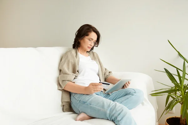 Middle-aged woman in casual clothes reading message, e-book or information on her tablet computer, sitting on white couch at home. Beautiful elderly female holding a credit card while shopping online