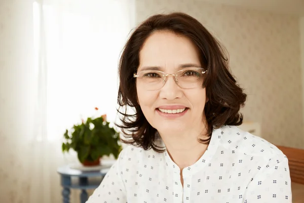 Portrait of beautiful elegant brunette middle-aged female looking with happy expression at the camera, sitting against cozy interior background, waiting for her children visiting her at the weekend