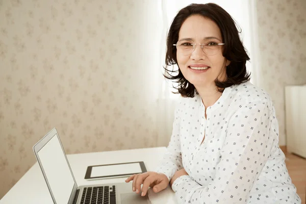 Close up shot of happy beautiful senior woman in glasses looking and smiling at the camera while typing a message or reading news on laptop with copy space screen for your text or promotional content